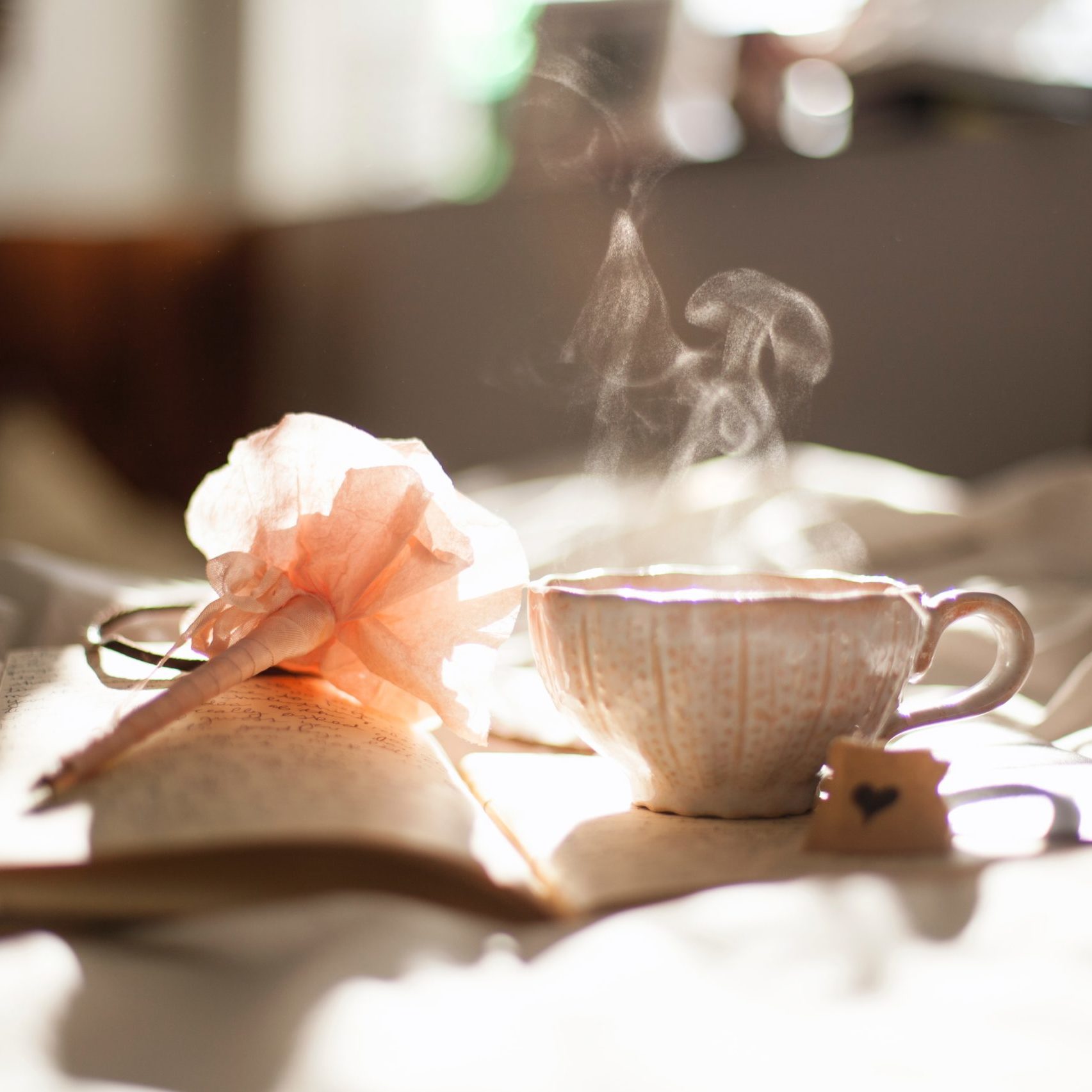 Teacup steaming in morning light on a notebook with written pages and with a pink flower-pen on top.