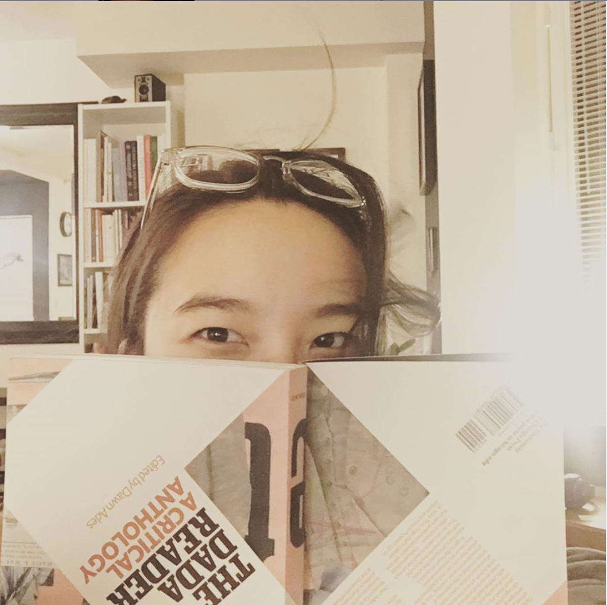 Ellen Chang-Richardson is hiding behind a book cover so you can only see their eyes and the top of their head with dark hair and glasses and a bookshelf in the background in this interior shot.