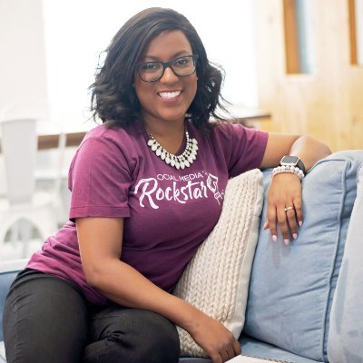 Andréa Jones wears a t-shirt that says Rockstar on it, a chunky necklace and glasses. She has medium-length black hair and sits on a blue and off-white cushioned couch.