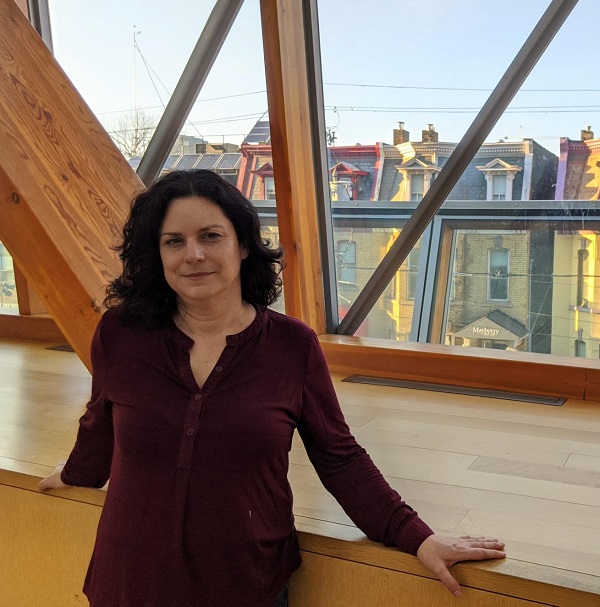 Lana has long dark wavy hair, wears a wine-coloured top, and is pictured before a big window with a wooden ledge overlooking row houses.