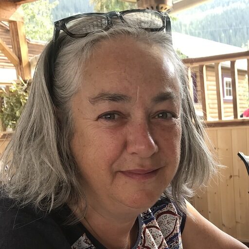 A smiling person with shoulder-length silver-gray hair sits on what appears to be a covered porch or deck. They are wearing sunglasses pushed up on their head and a patterned sleeveless top. Behind them, wooden railings and cabin-style buildings are visible with mountain scenery in the background.