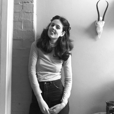 Lyndall Cain stands against a brick and smooth wall, wearing long hair pinned back, black jeans and a long-sleeved turtleneck shirt. There is a small skull with antlers on the wall to Lyndall’s left..