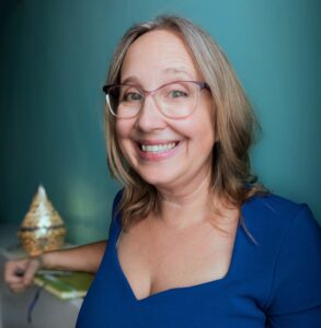 Rachel Thompson has yellow and grey hair and wears glasses and a blue dress with a green notebook and teal wall behind her in this portrait.