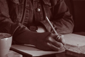 A hand writing in a notebook with a cup on a table. The person wears a jean jacket.