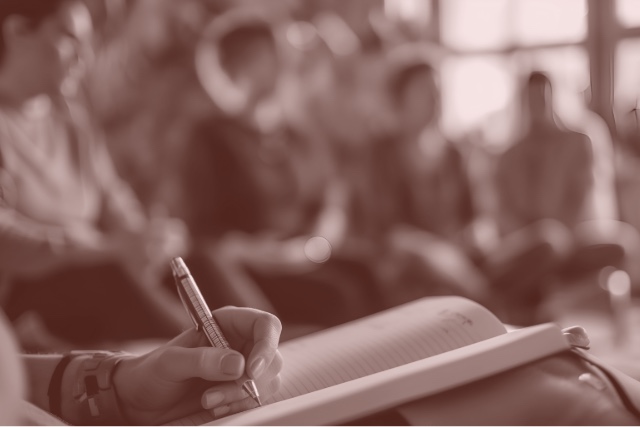 A group of writers sitting in a circle with a closeup of one writer's hand on a pen in an open notebook.