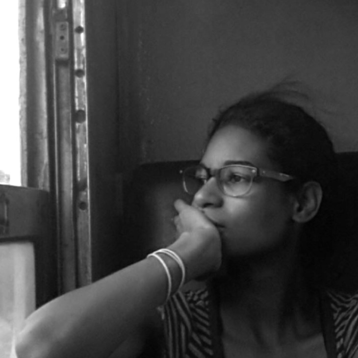 The image is a black-and-white photograph of a person sitting by a window, looking thoughtfully outside. They are wearing rectangular glasses, and have their hair pulled back. Their hand rests against their face, partially covering their mouth, and they're wearing a light-colored bracelet on their wrist. The overall mood of the image feels contemplative and introspective.