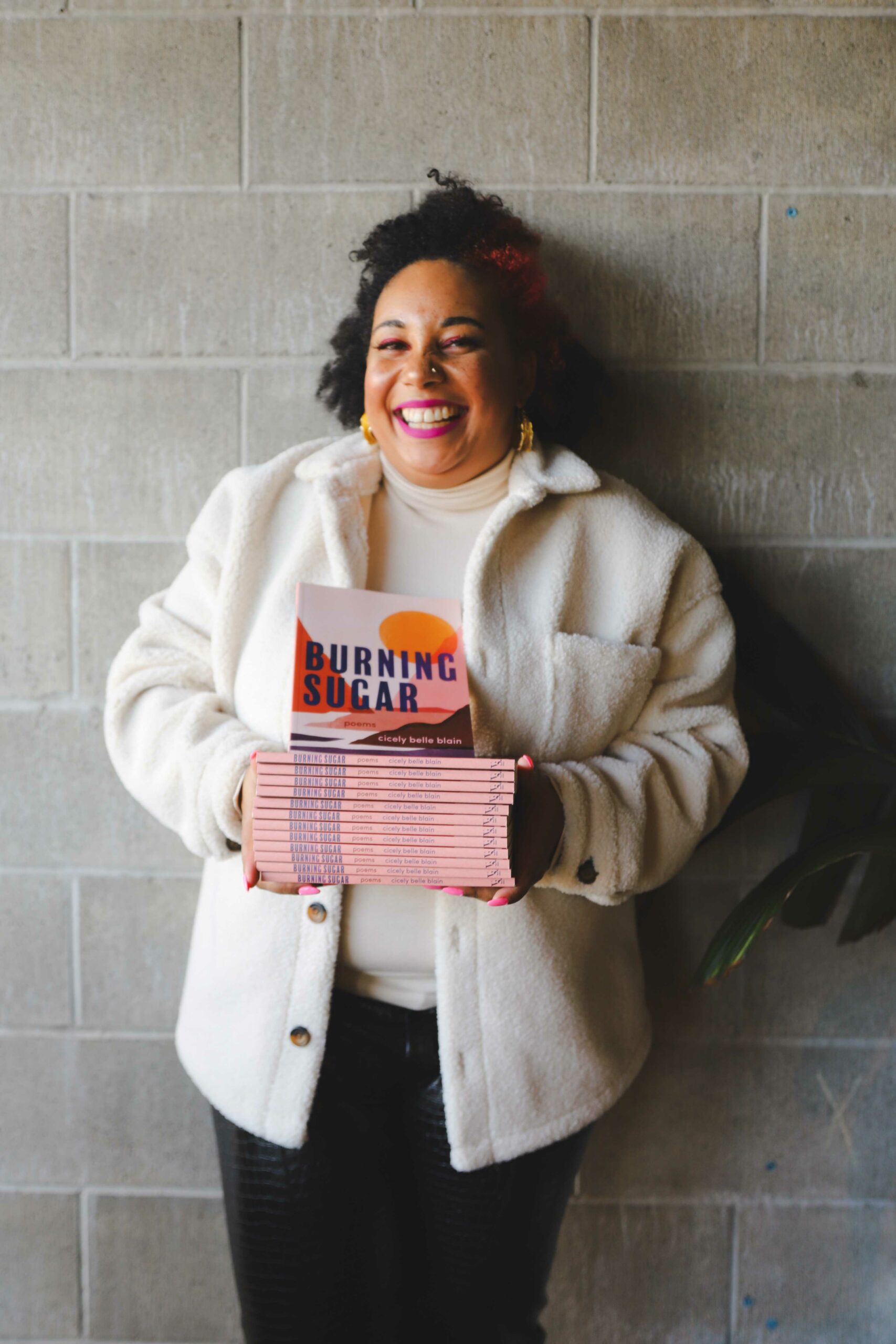 Cicely Belle Blain is photographed with a stack of their book of poetry, Burning Sugar, and smiling widely in front of a brick wall and wearing a white jacket and turtleneck with gold earrings and mid-length curly haired half tied up.