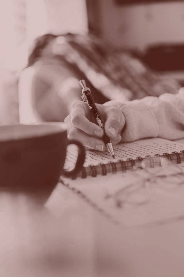 A woman in a yellow sweater shirt sits in a chair and writes in a planner.