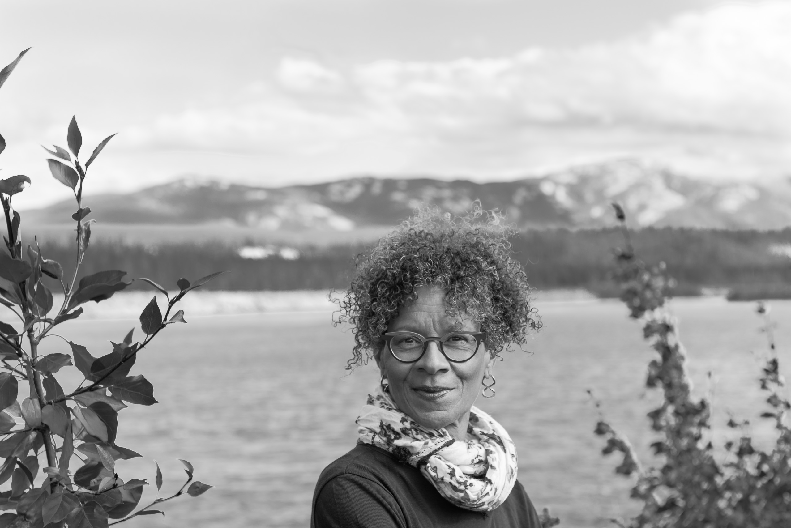 Tina Brobby has curly hair, chunky glasses, earrings, and a scarf in this black and white photo taken outdoors before a body of water with mountains in the background and foliage around the frame.